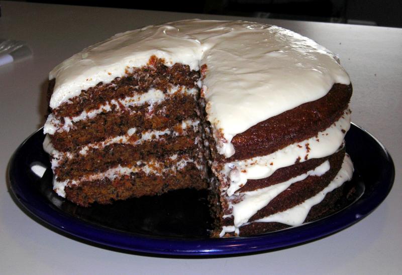 A round four-layer carrot cake on a blue plate.  A wedge account for about a fifth of the cake has been cut away, showing the internal layer structure.